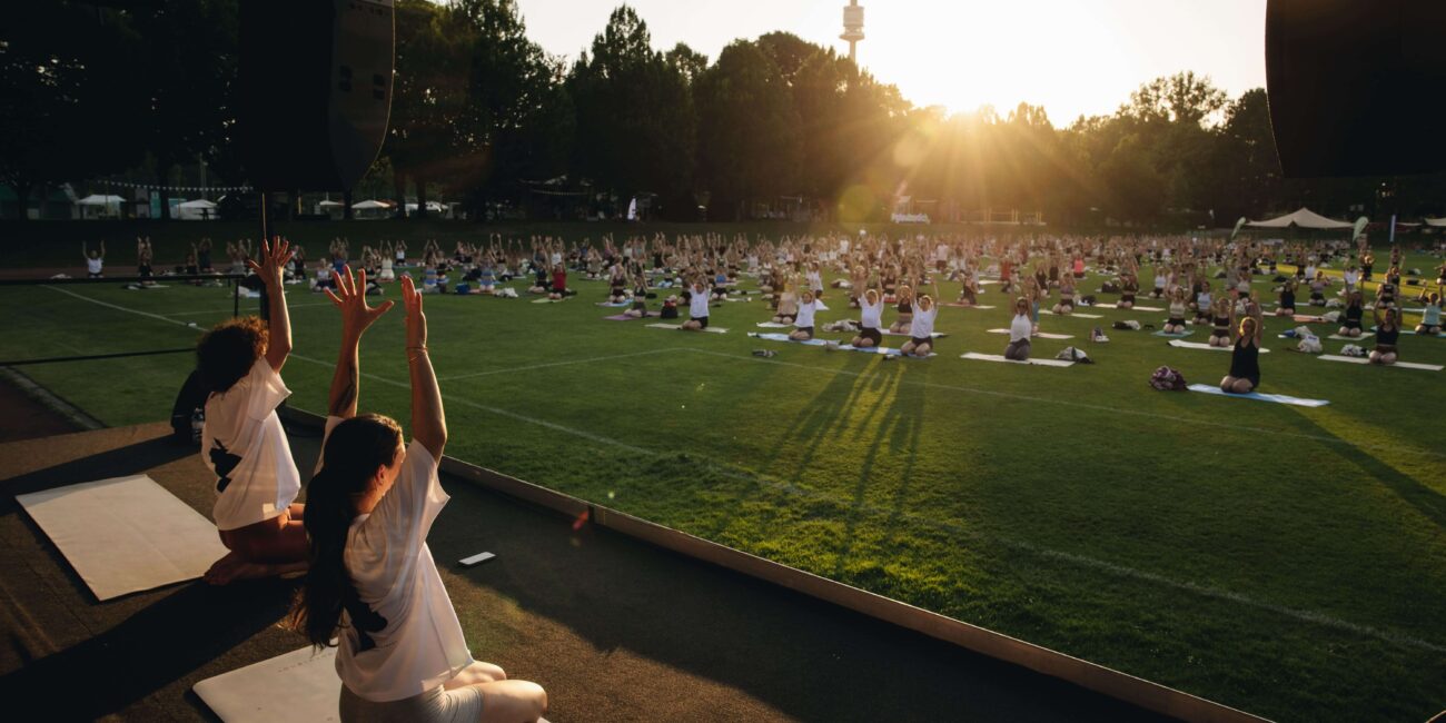 Yogalehrerinnen knien mit erhobenen Armen auf der Bühne vor der Gruppe bei goldenem Abendlicht