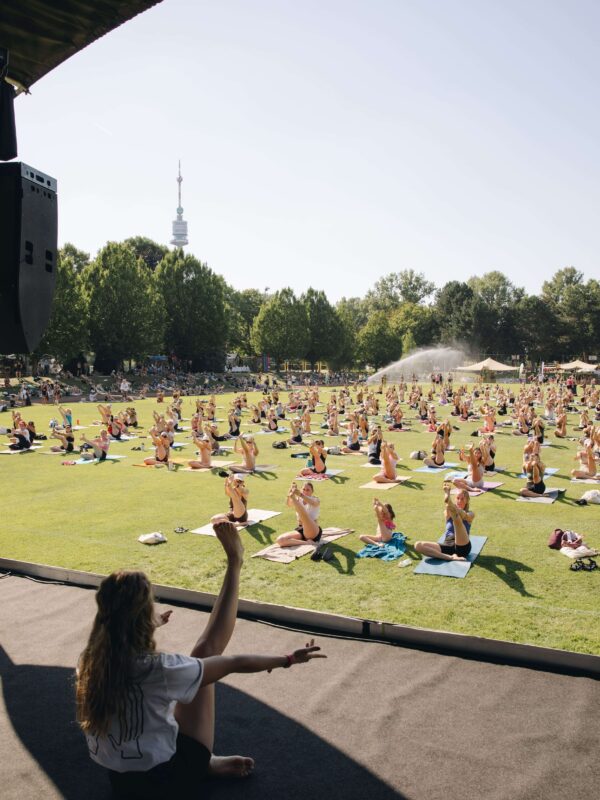 Yogalehrerin gestikuliert von der Bühne, während eine große Gruppe auf der Wiese Yoga übt