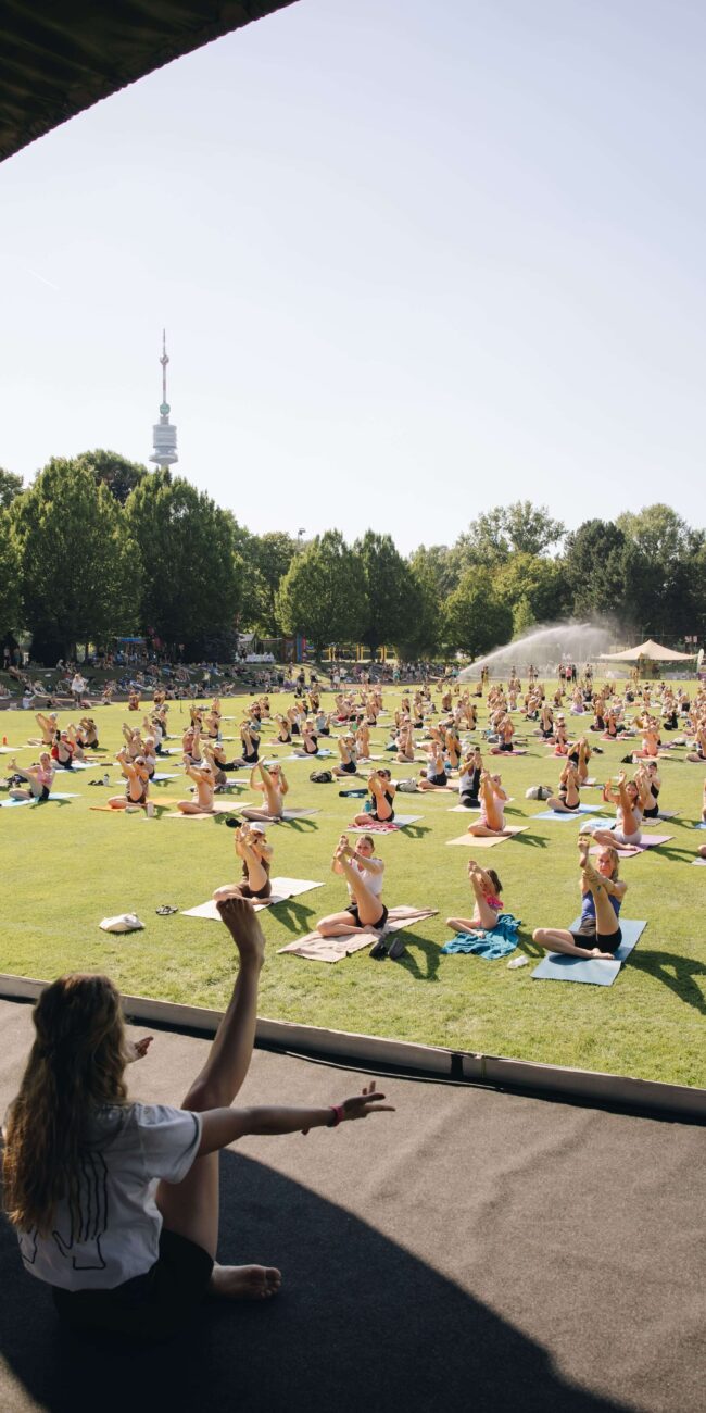 Yogalehrerin gestikuliert von der Bühne, während eine große Gruppe auf der Wiese Yoga übt